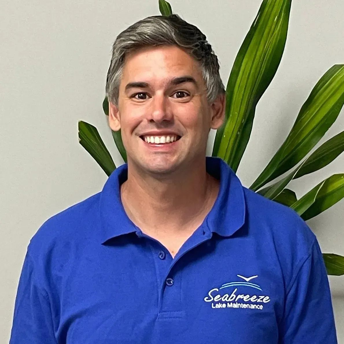 Smiling man in a blue polo shirt standing indoors with green plant leaves behind him