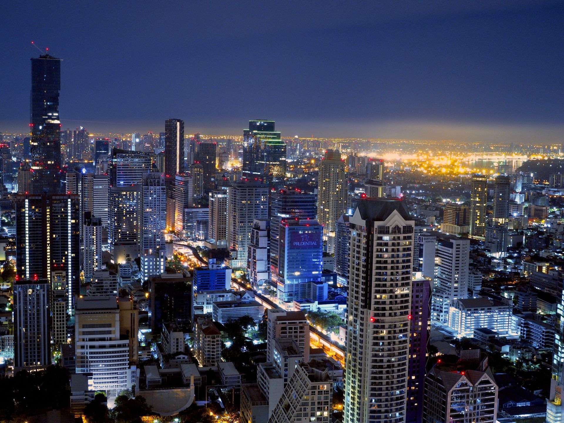 City skyline at night, numerous skyscrapers lit up against a dark blue sky, with a hazy glow on the horizon.