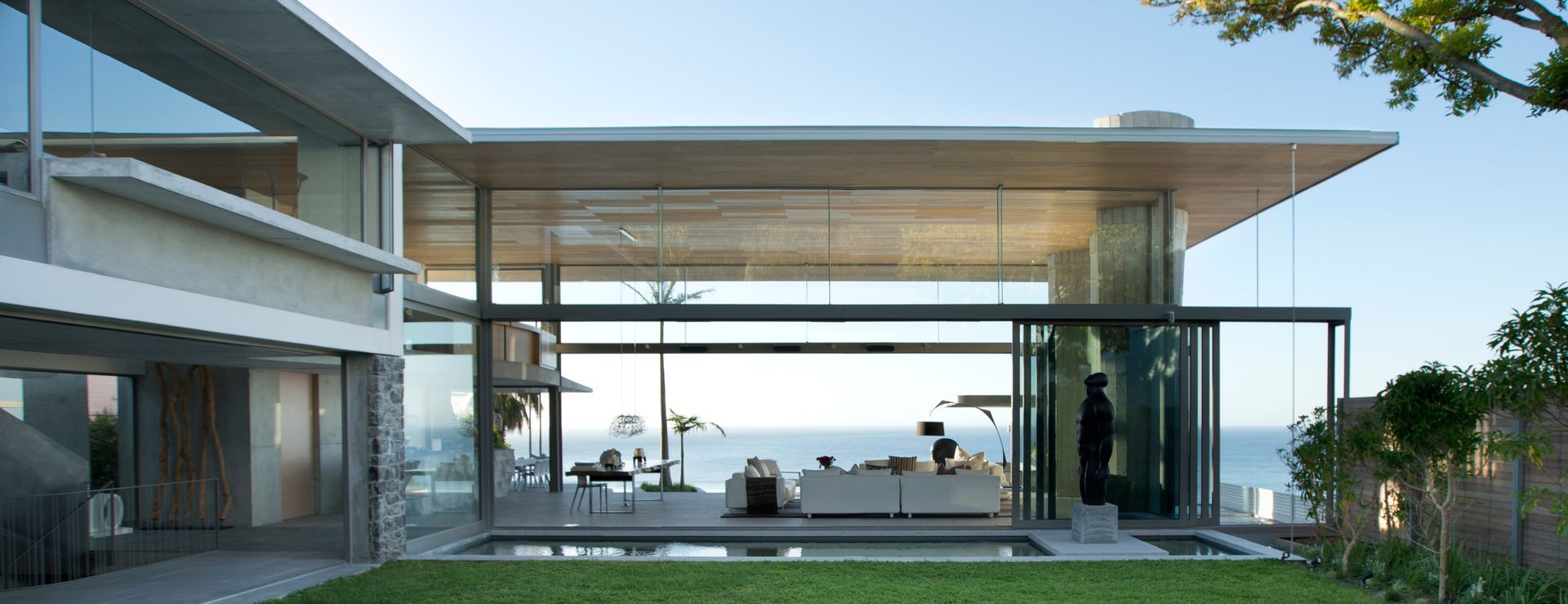 Modern house with glass walls overlooks a pool and ocean.