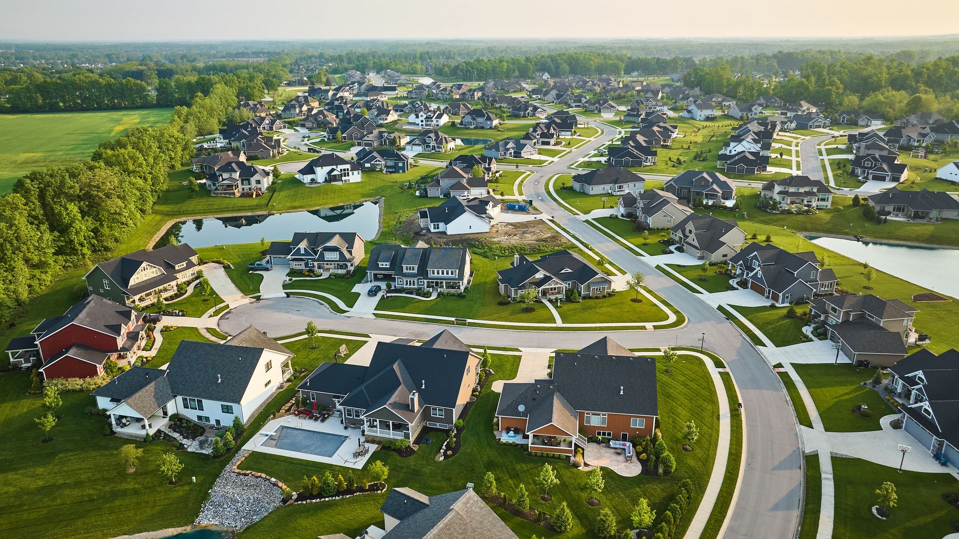 Aerial view of a suburban neighborhood with houses, roads, ponds, and greenery under a blue sky.