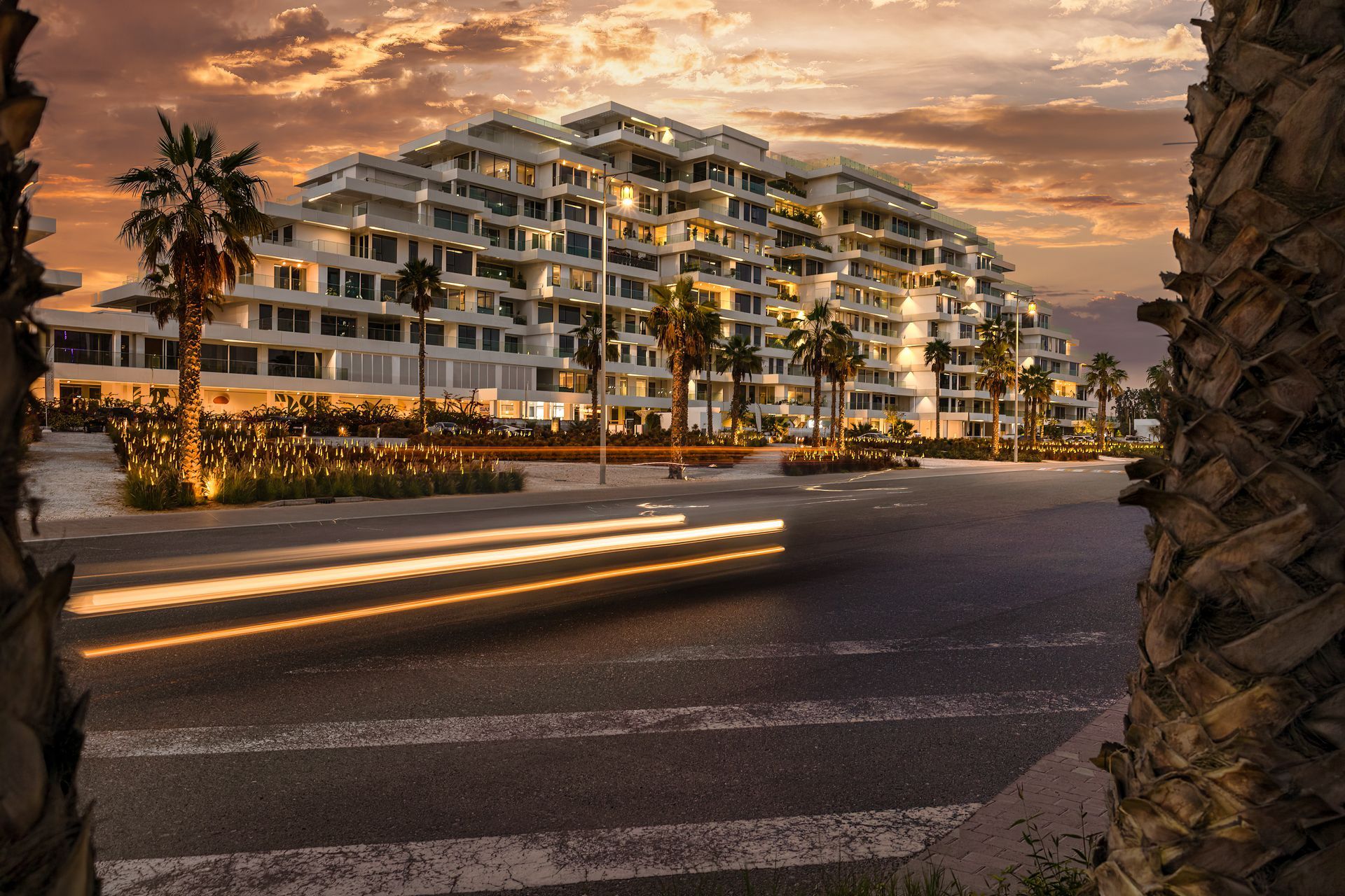 Modern terraced building with lights, palm trees, and blurred car lights on a road at sunset.
