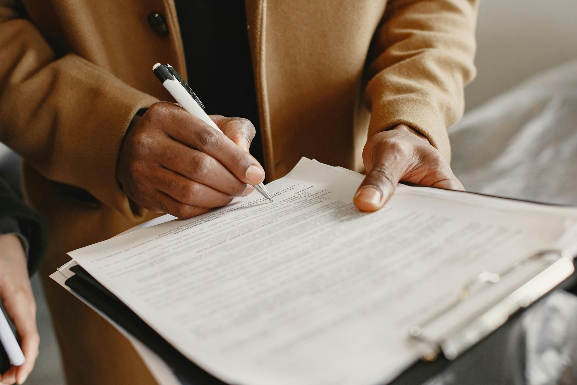 Hands signing a document on a clipboard with a pen in a tan coat sleeve