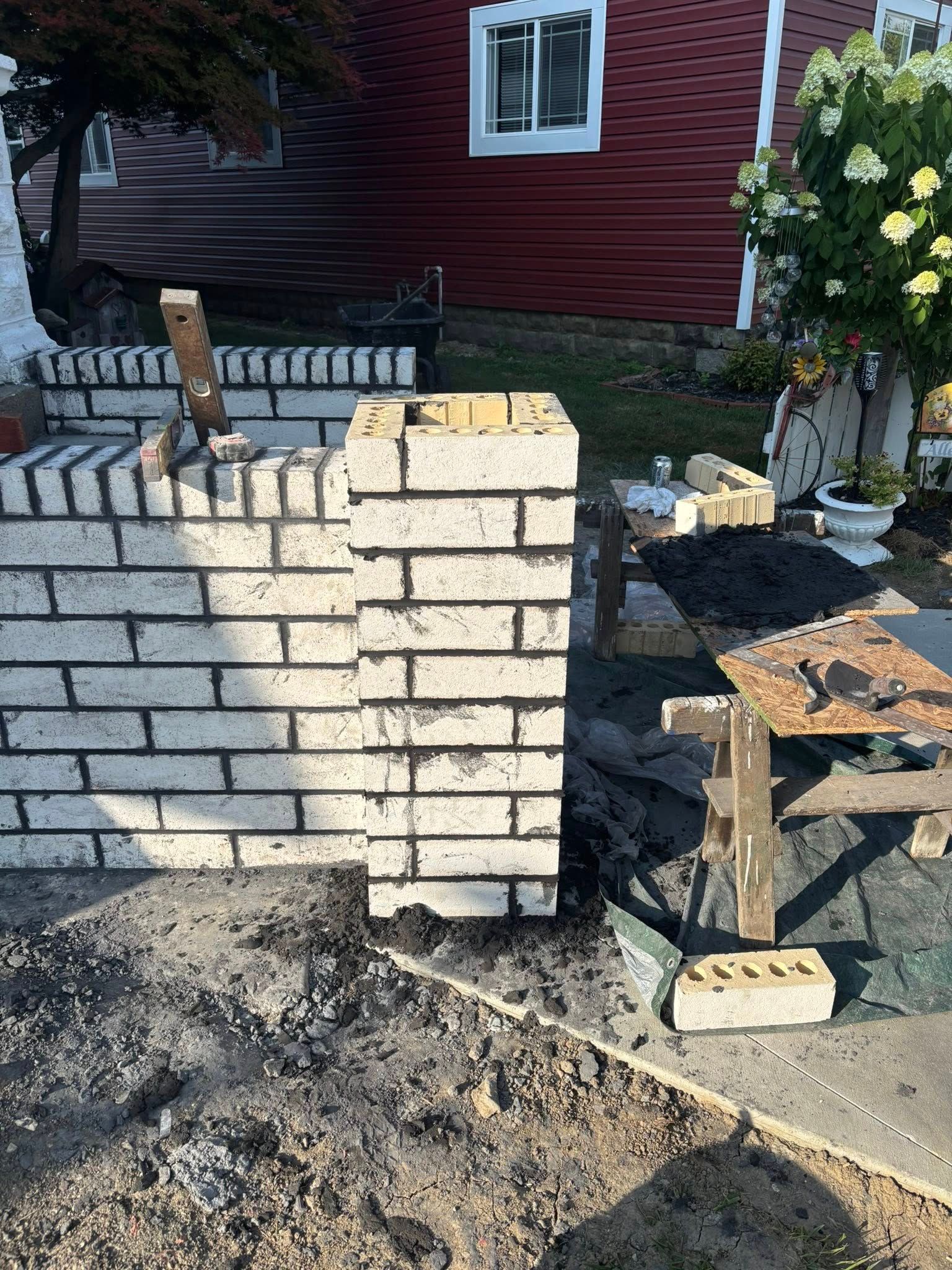 Brick pillars and wall under construction outdoors with red house in background.