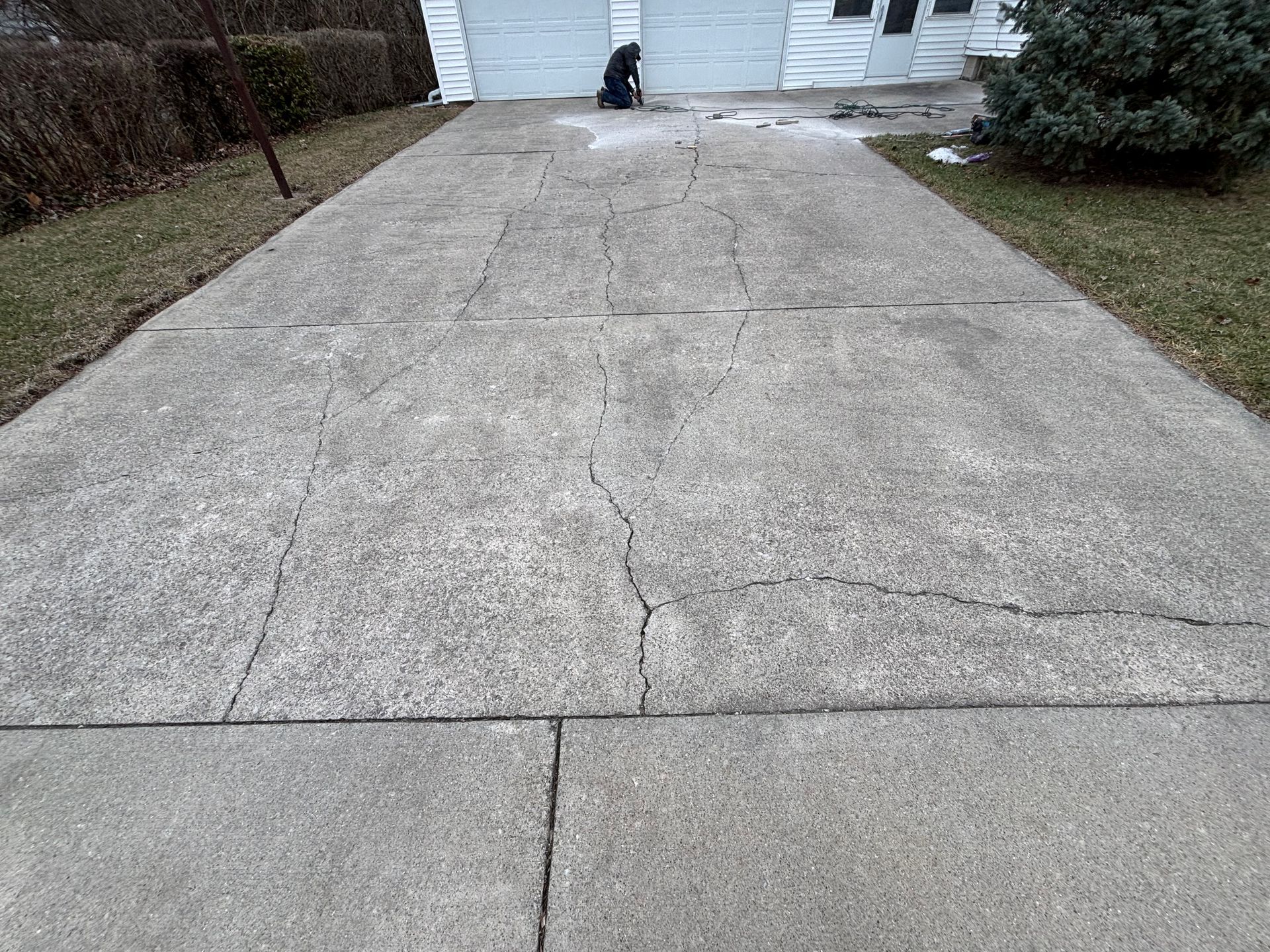 Cracked concrete driveway leading to a white garage, flanked by grass and shrubbery.