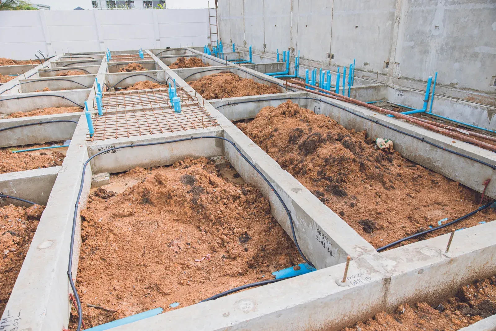 Concrete rectangular beds with dirt, plumbing, and blue pipes; outdoor construction site.
