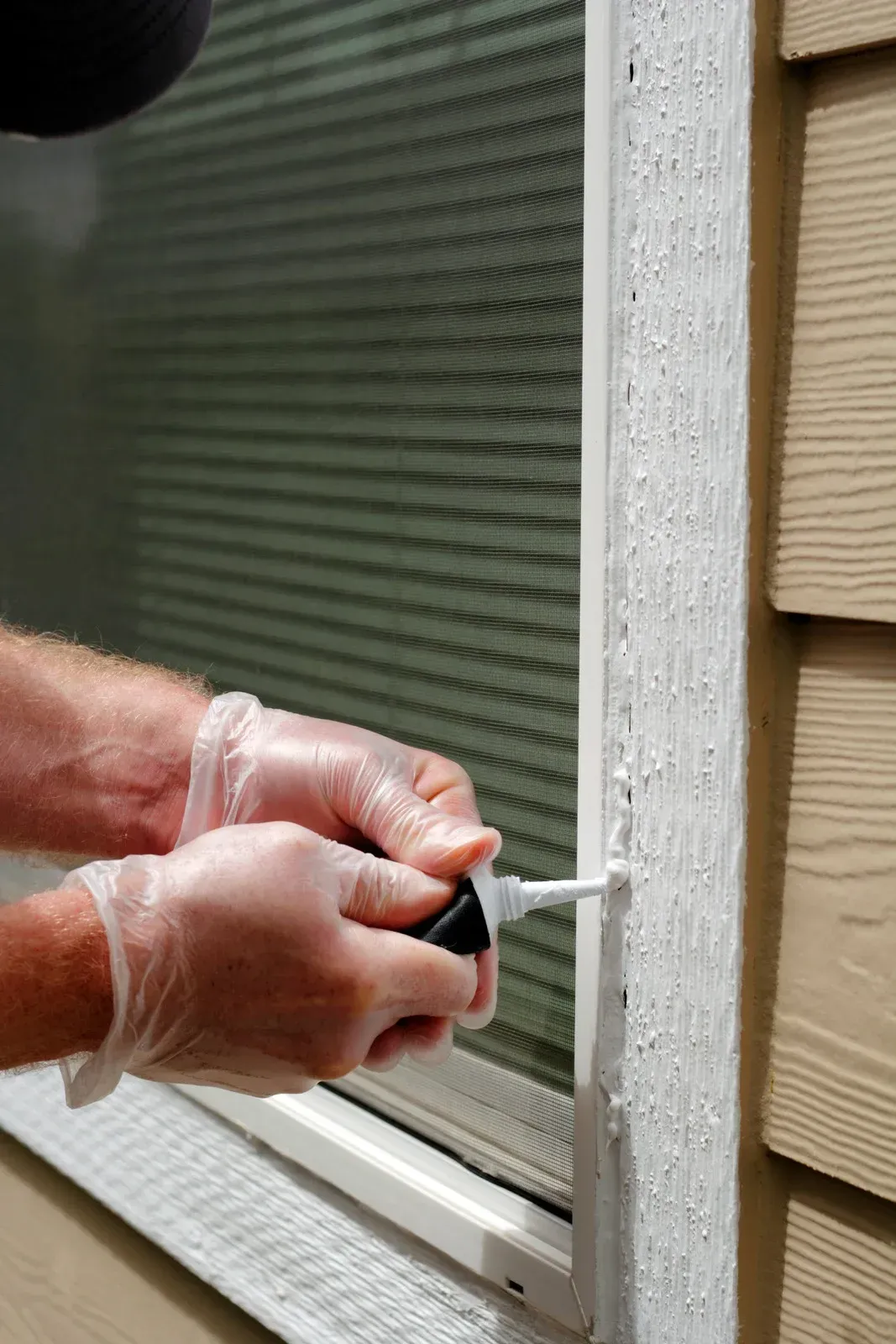 Person applying caulk to a window frame, wearing gloves, outdoors.