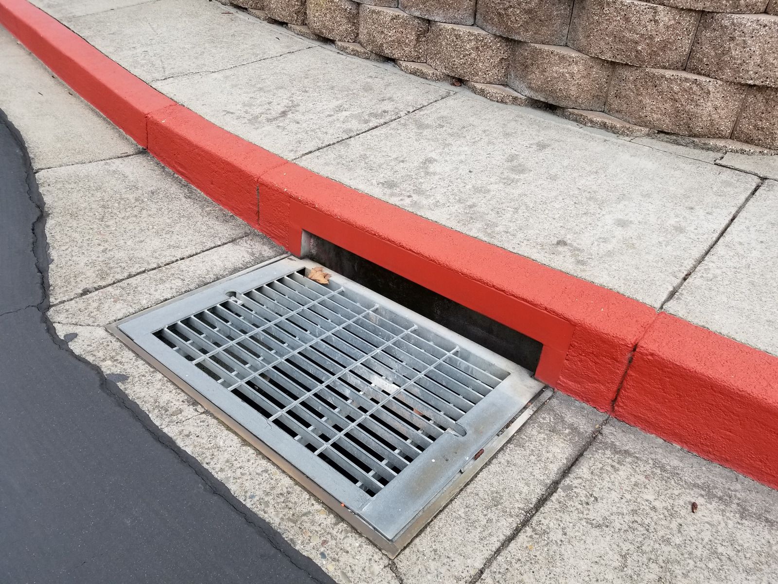 Metal grate drain in red painted curb, on a sidewalk, next to a brick wall.