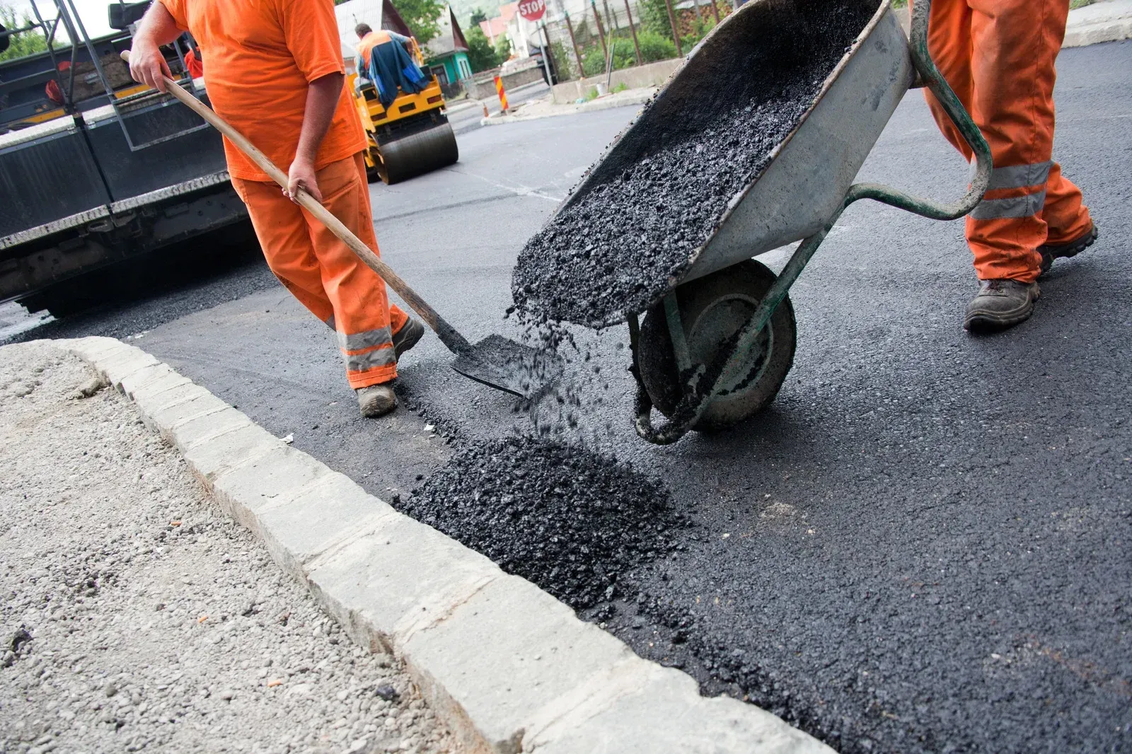 Road workers laying asphalt, using shovels and a wheelbarrow. Orange work suits.