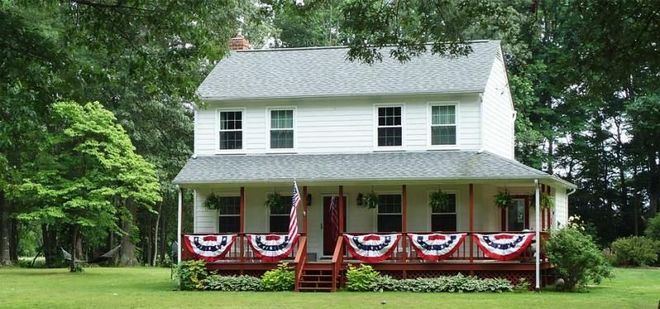 White farmhouse with red porch bunting amid green trees and lawn