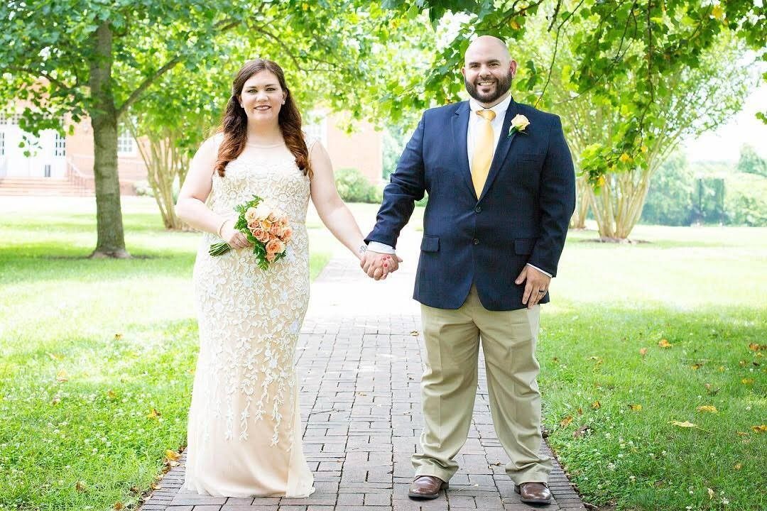 Couple holding hands on a garden path, bride in white dress and groom in navy suit, smiling outdoors.
