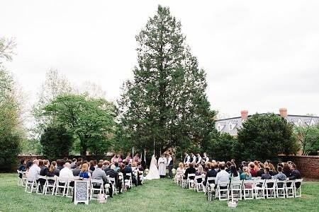 Outdoor wedding ceremony with guests seated on white chairs around a large tree on a grassy lawn
