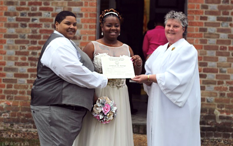 Three people holding a certificate at an outdoor ceremony, standing before a brick wall.