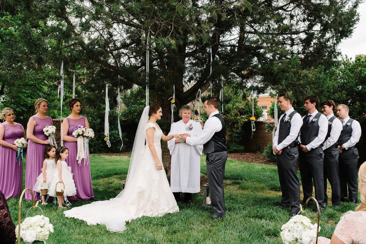 Outdoor wedding ceremony with bride, groom, officiant, bridesmaids, and groomsmen under trees