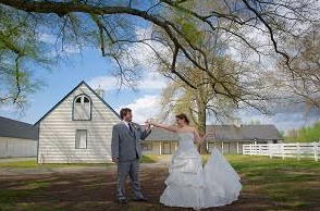 Bride and groom dancing outdoors beside a white barn under a large tree.