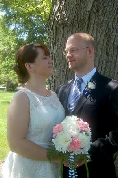 Wedding couple standing by a tree, smiling and holding a pink-and-white bouquet outdoors.