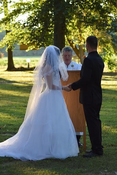 Bride and groom stand with an officiant outdoors under a tree during a wedding ceremony.