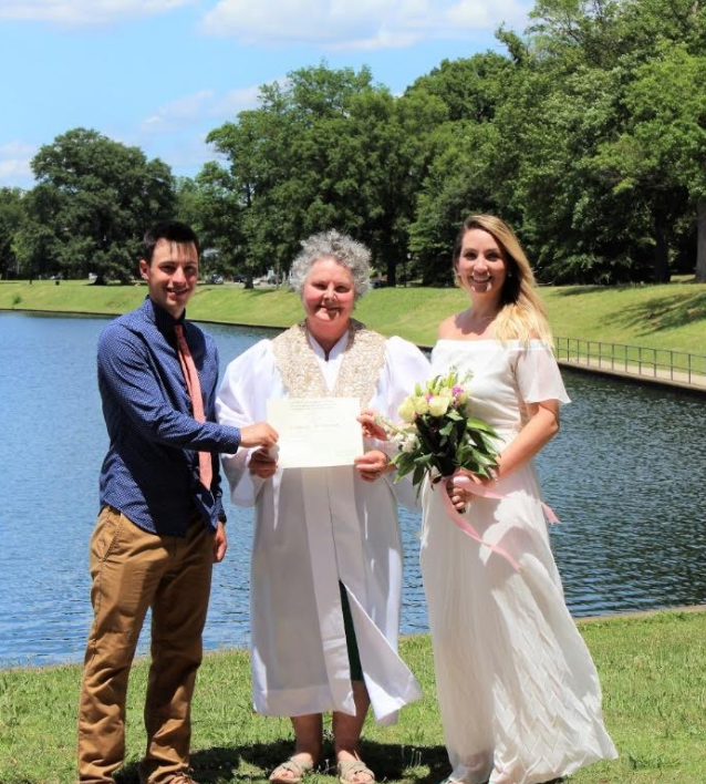Three people stand by a lake holding a certificate and bouquet at an outdoor wedding ceremony.