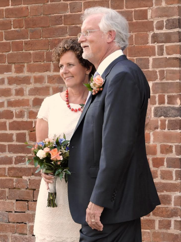 Couple in formal attire standing together by a brick wall, holding a bouquet and smiling.