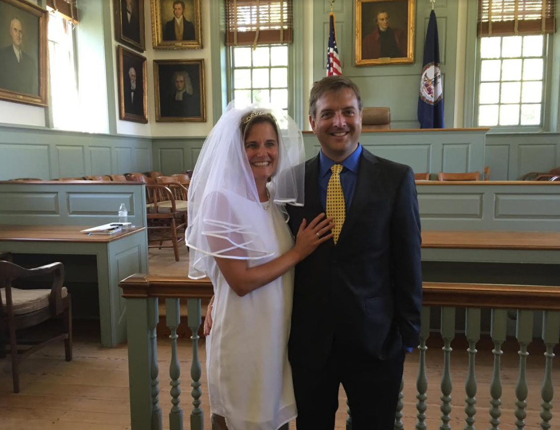Bride and groom pose smiling in a courthouse ceremony room, with windows and portraits behind them.