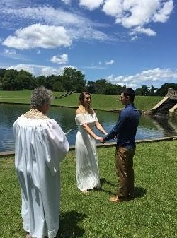 Two people holding hands by a lake during an outdoor wedding ceremony, with an officiant nearby.