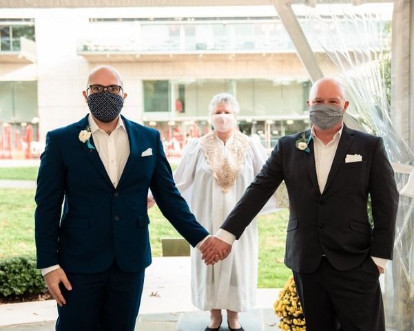 Three masked people standing together, with two men holding hands in front of a bright outdoor entrance.