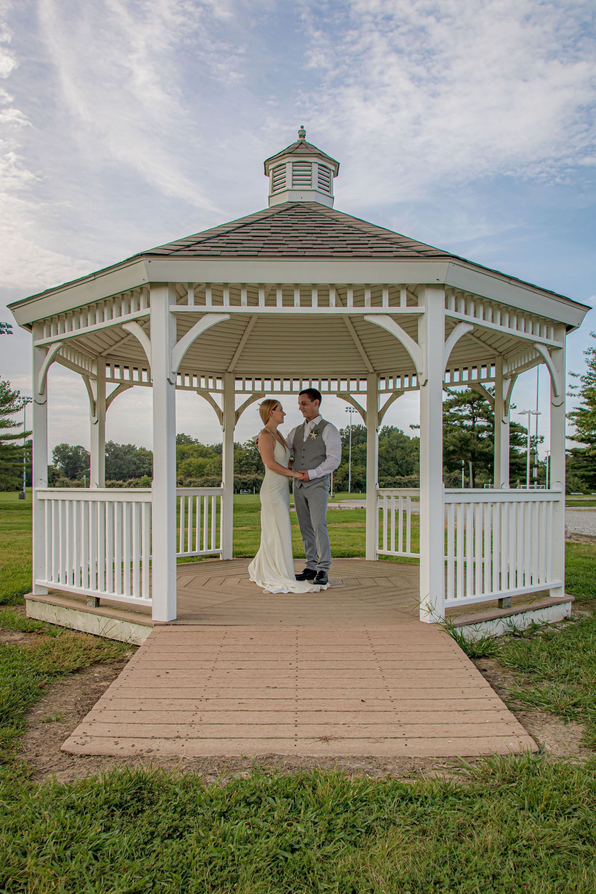 Couple in wedding attire standing in a white gazebo on a grassy lawn under a cloudy sky