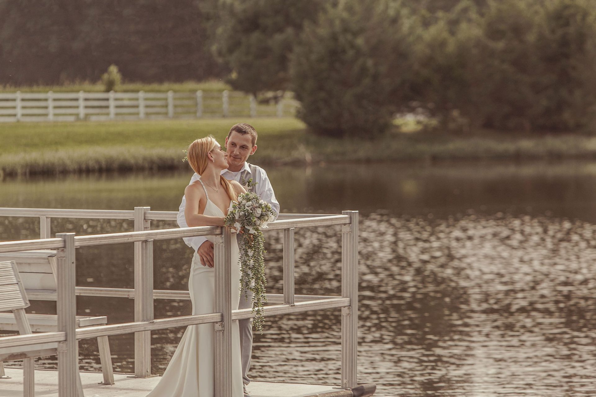 Couple in wedding attire standing on a dock by a calm lake, embracing outdoors.