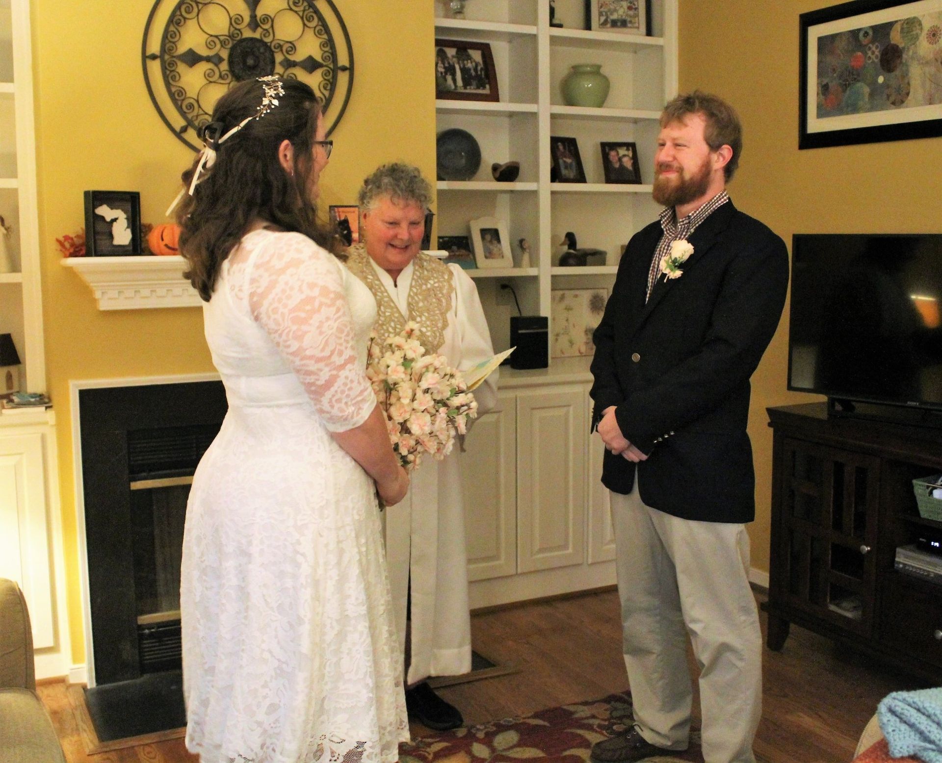 Three people stand in a cozy living room during a wedding ceremony, with the bride in white and groom in a dark jacket.