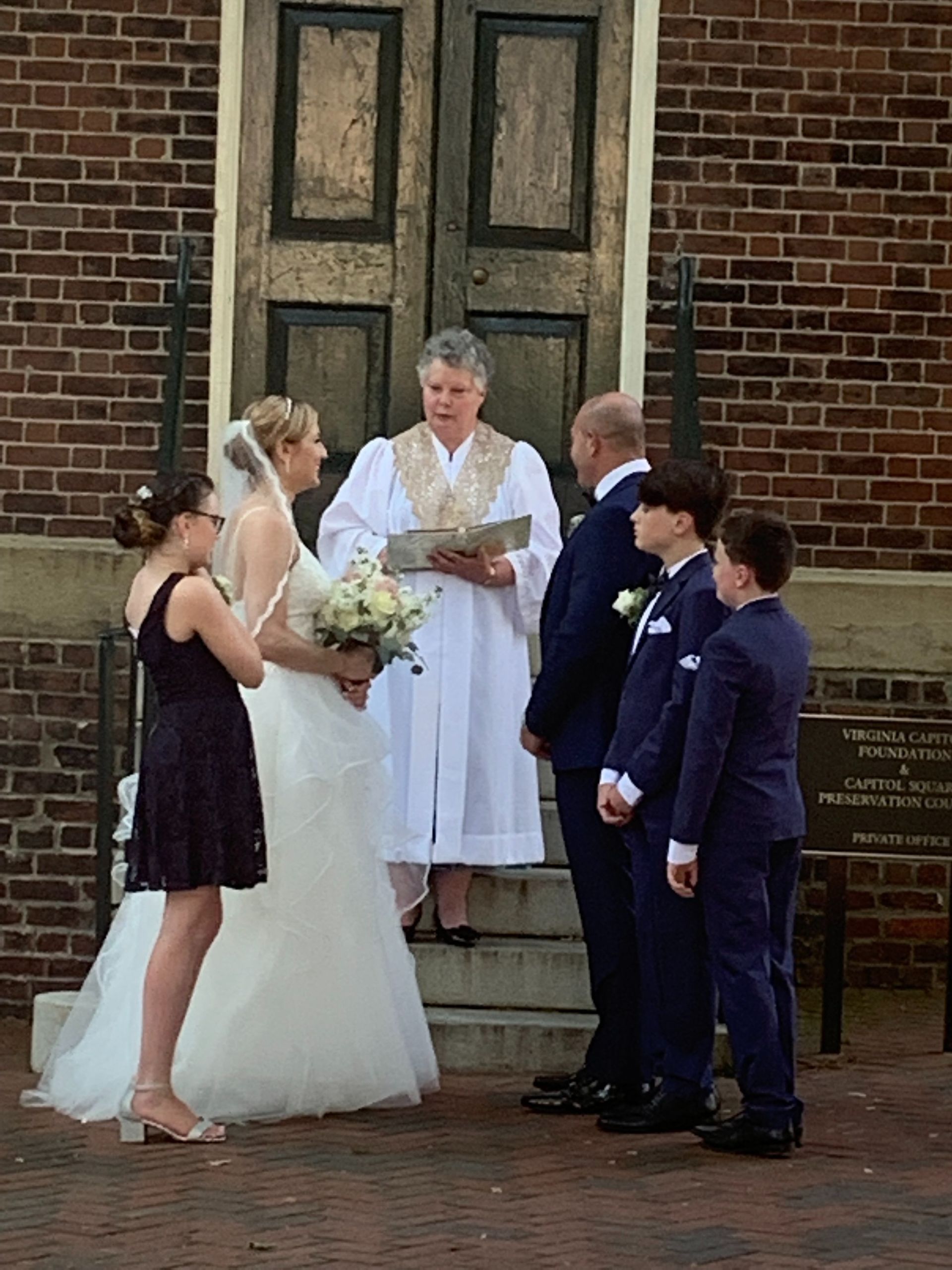 Outdoor wedding ceremony with bride, groom, officiant, and attendants outside a brick building
