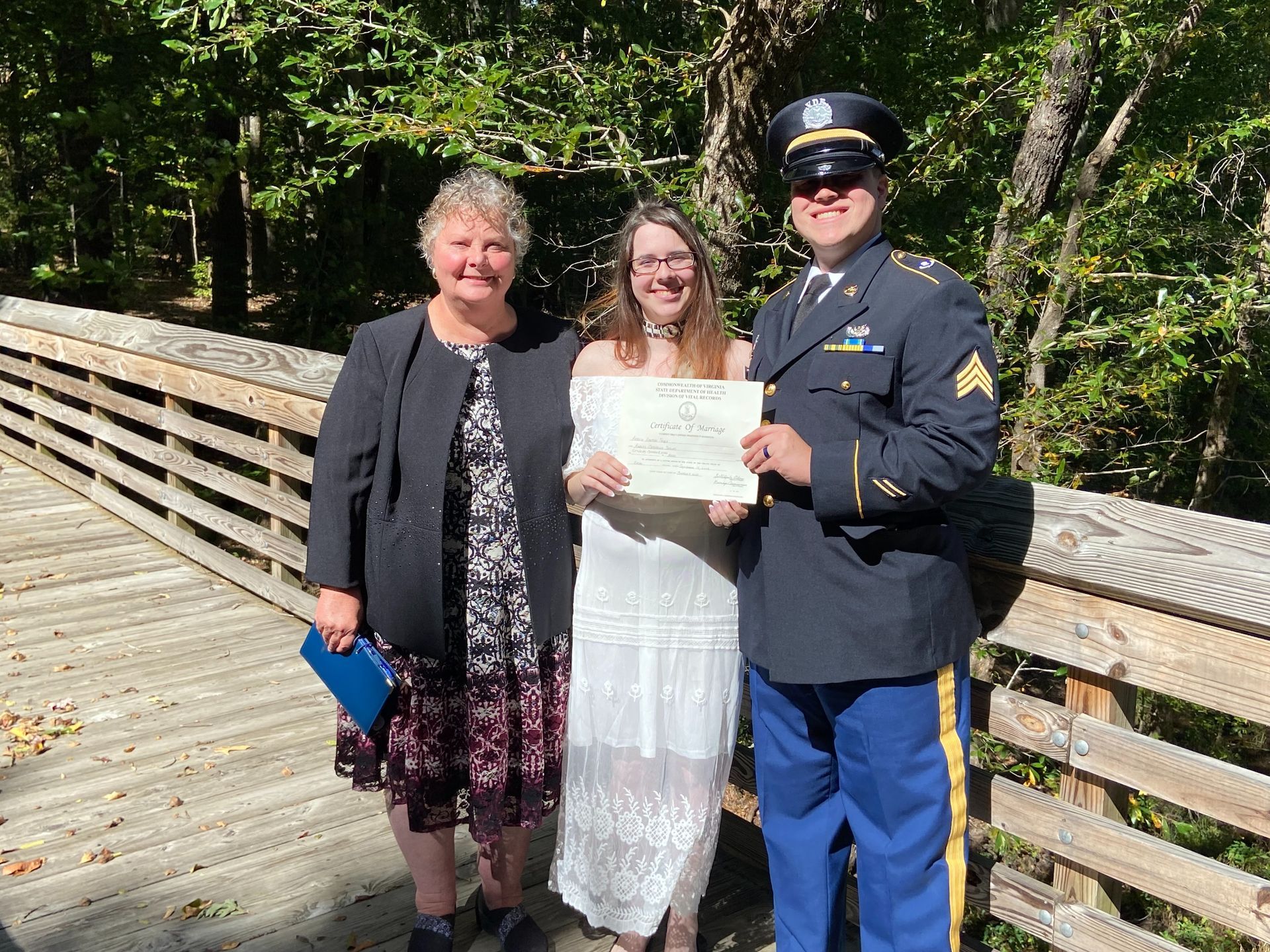 Three people on a wooden bridge, two holding a certificate, one in a military uniform, in a sunny wooded area