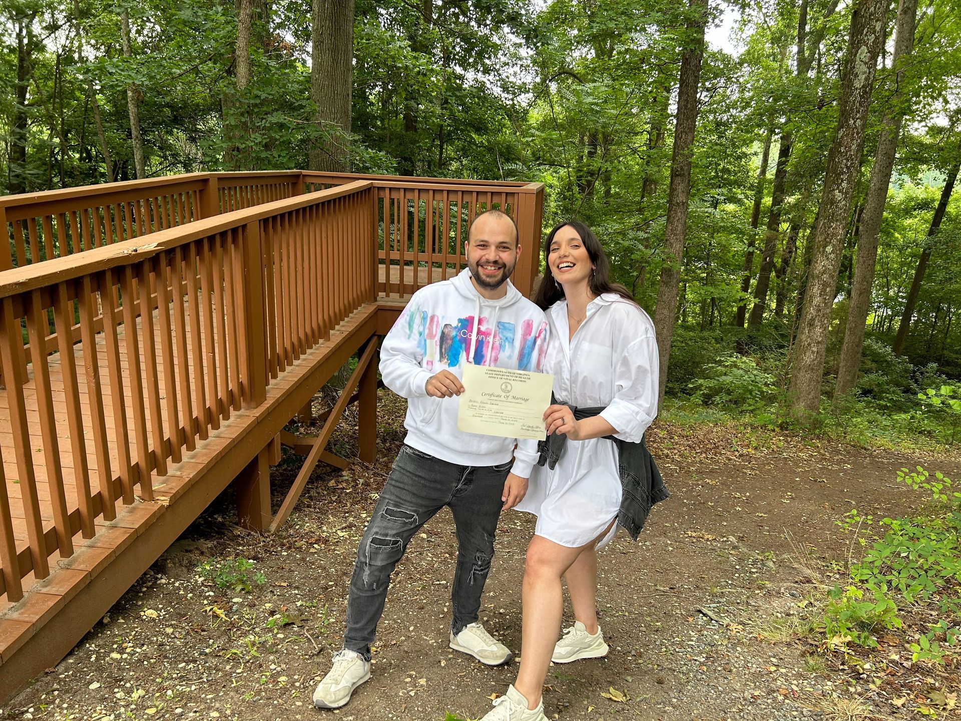 Two people smile on a wooded trail, holding a map beside a wooden boardwalk.