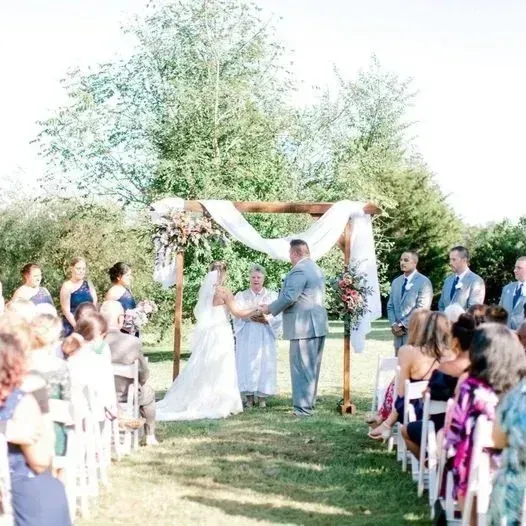 Outdoor wedding ceremony under a white draped arch with guests seated on both sides of an aisle.