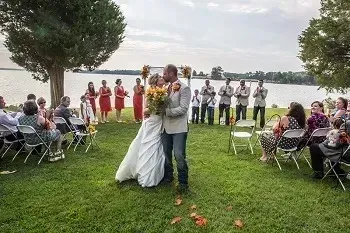 Outdoor wedding couple kissing on a grassy lawn by the lake, with guests seated and wedding party standing nearby