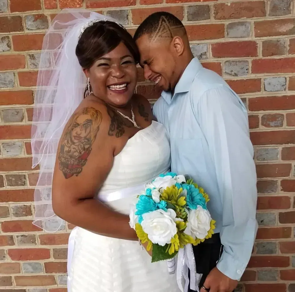 Smiling bride and groom embracing, holding blue and white bouquet, standing before a brick wall.