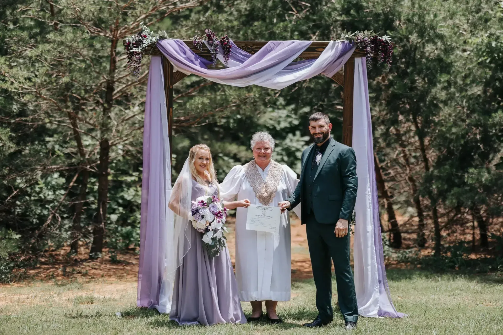 Three people posing under a purple-draped arch in a wooded outdoor setting, holding a bouquet.