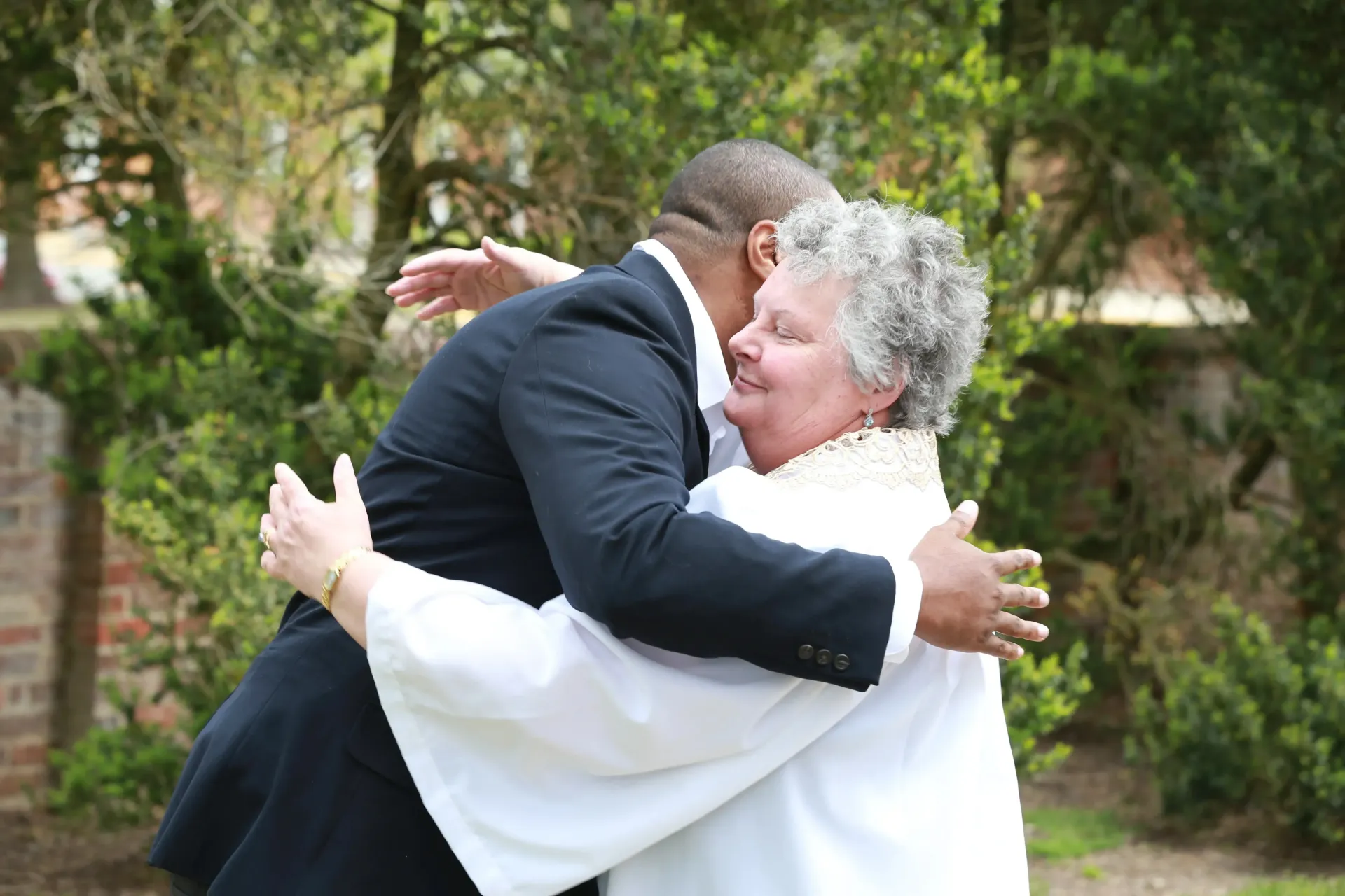 Two people hugging outdoors, one in a dark suit and the other in a white jacket, with trees in the background.