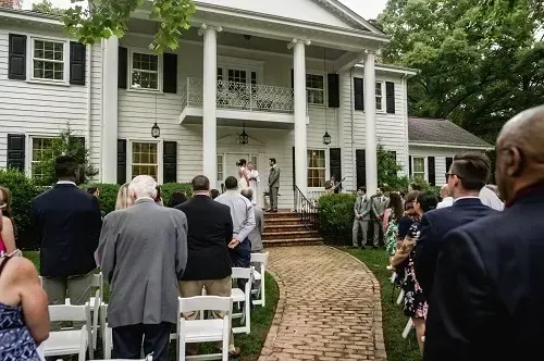Outdoor wedding ceremony in front of a white house with guests seated on a brick path