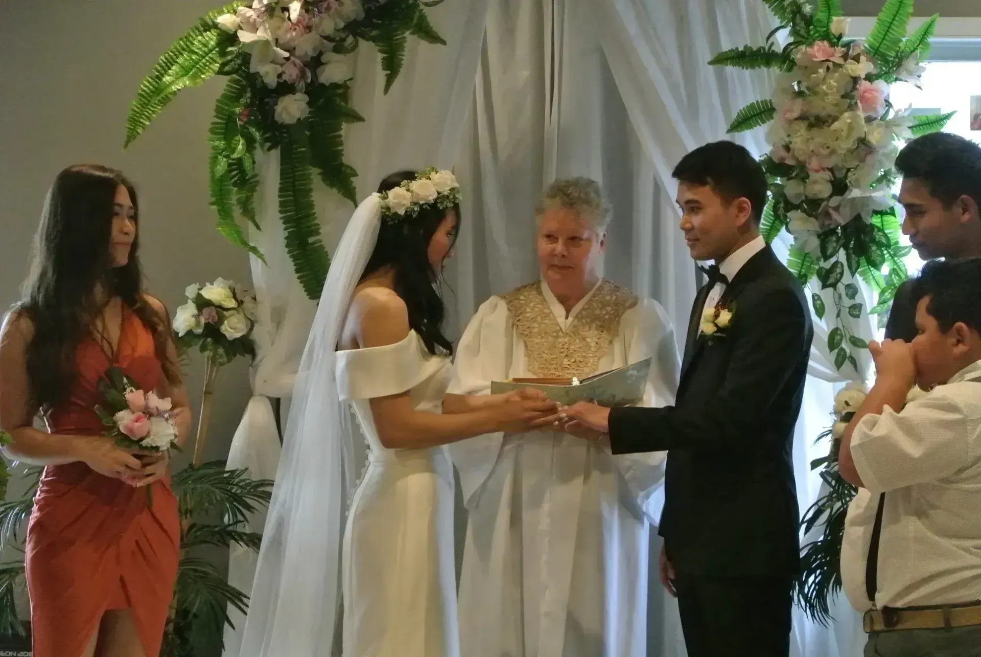 Wedding ceremony with bride and groom exchanging rings beside an officiant, under floral arch.
