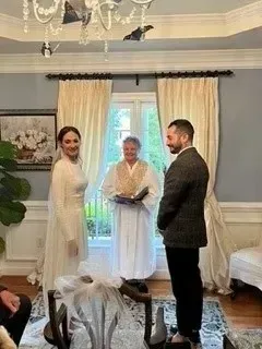 Three people stand in a bright living room during an indoor ceremony, with a window and curtains behind them.