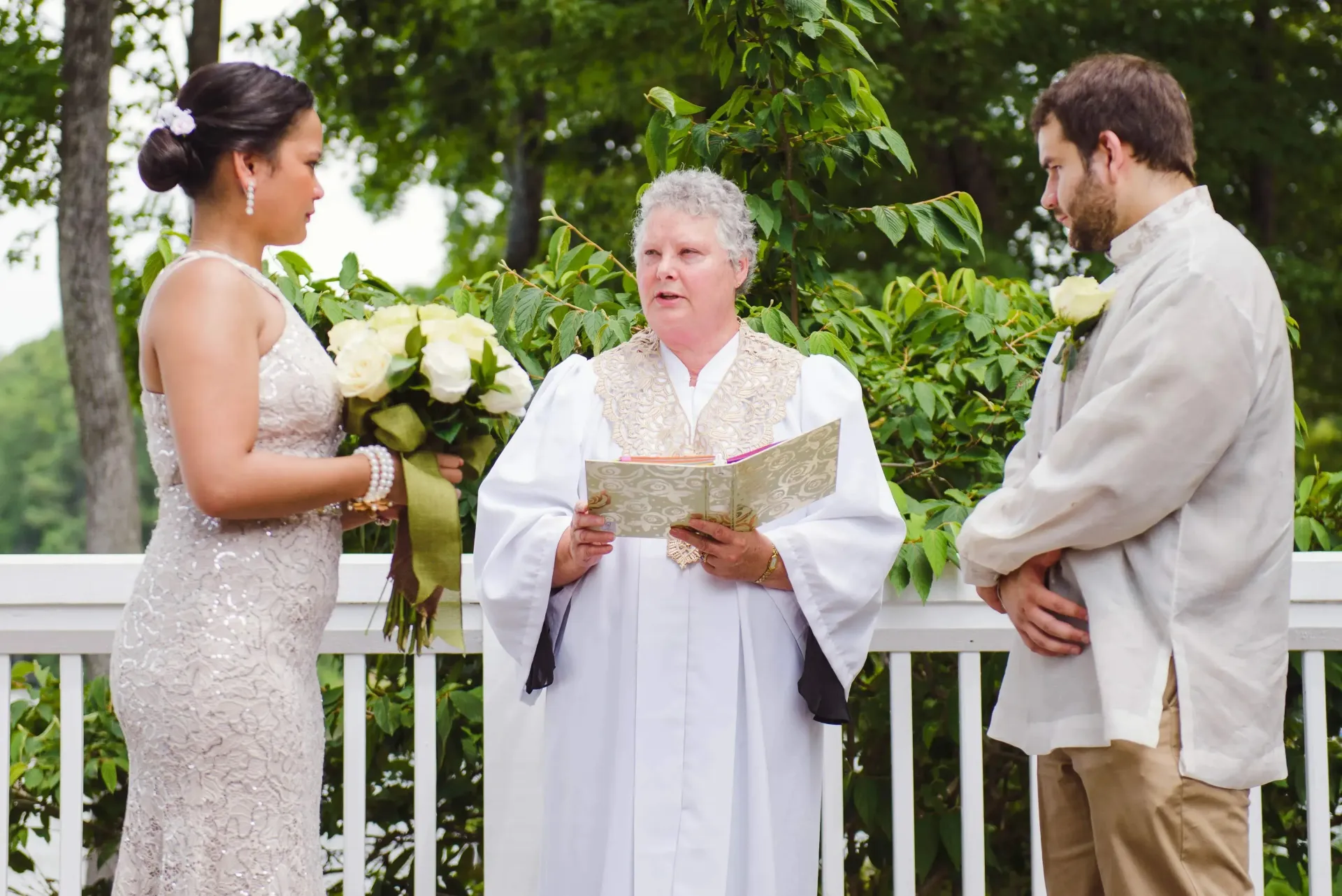 Wedding ceremony outdoors with officiant, bride in white dress, and groom facing each other