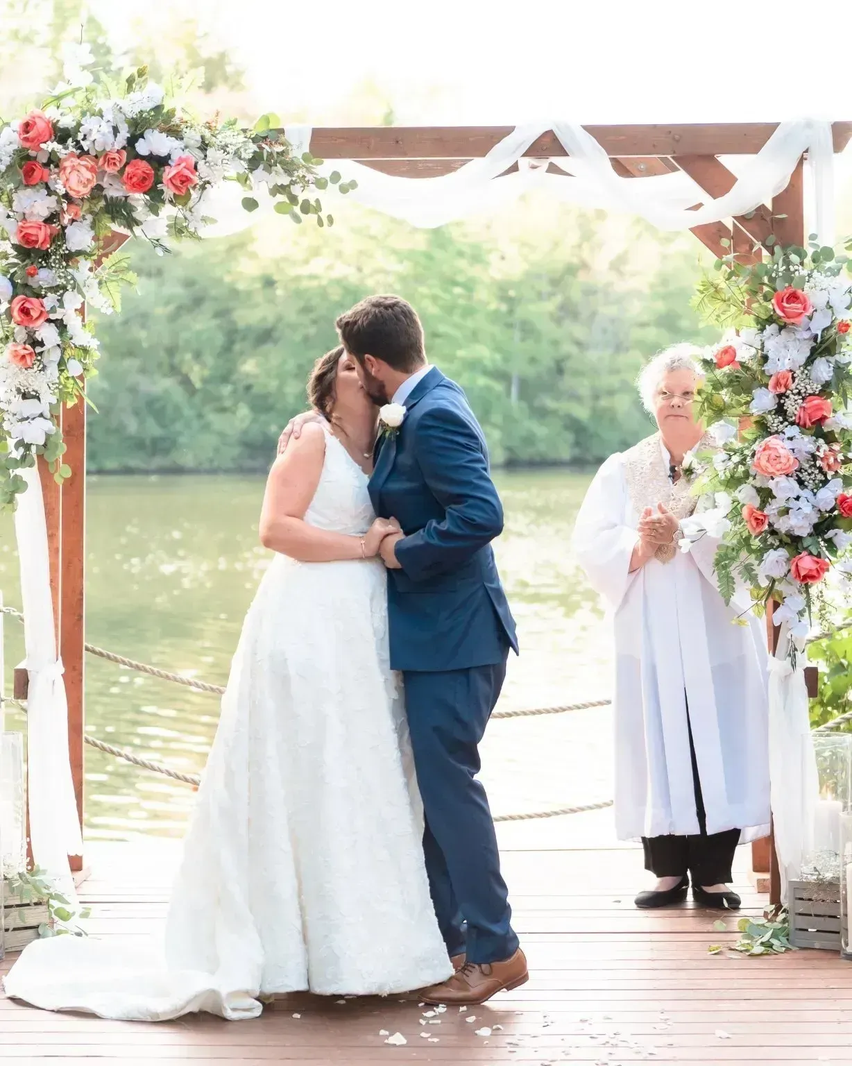 Bride and groom kissing beneath a floral arch on a lakeside dock at an outdoor wedding