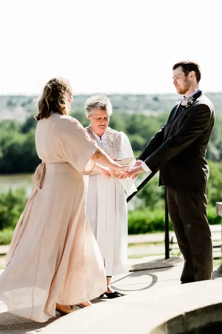 Outdoor wedding ceremony with two people holding hands before an officiant, in bright sunlight.