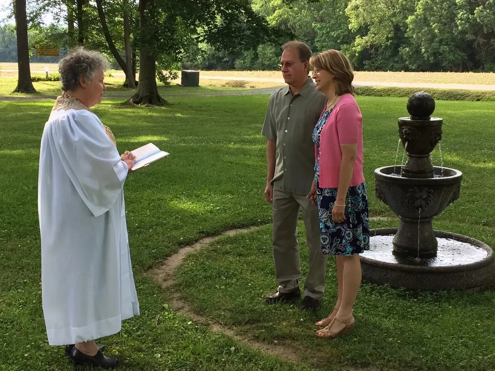 Two people stand by a garden fountain while a person in white robes reads from a paper.