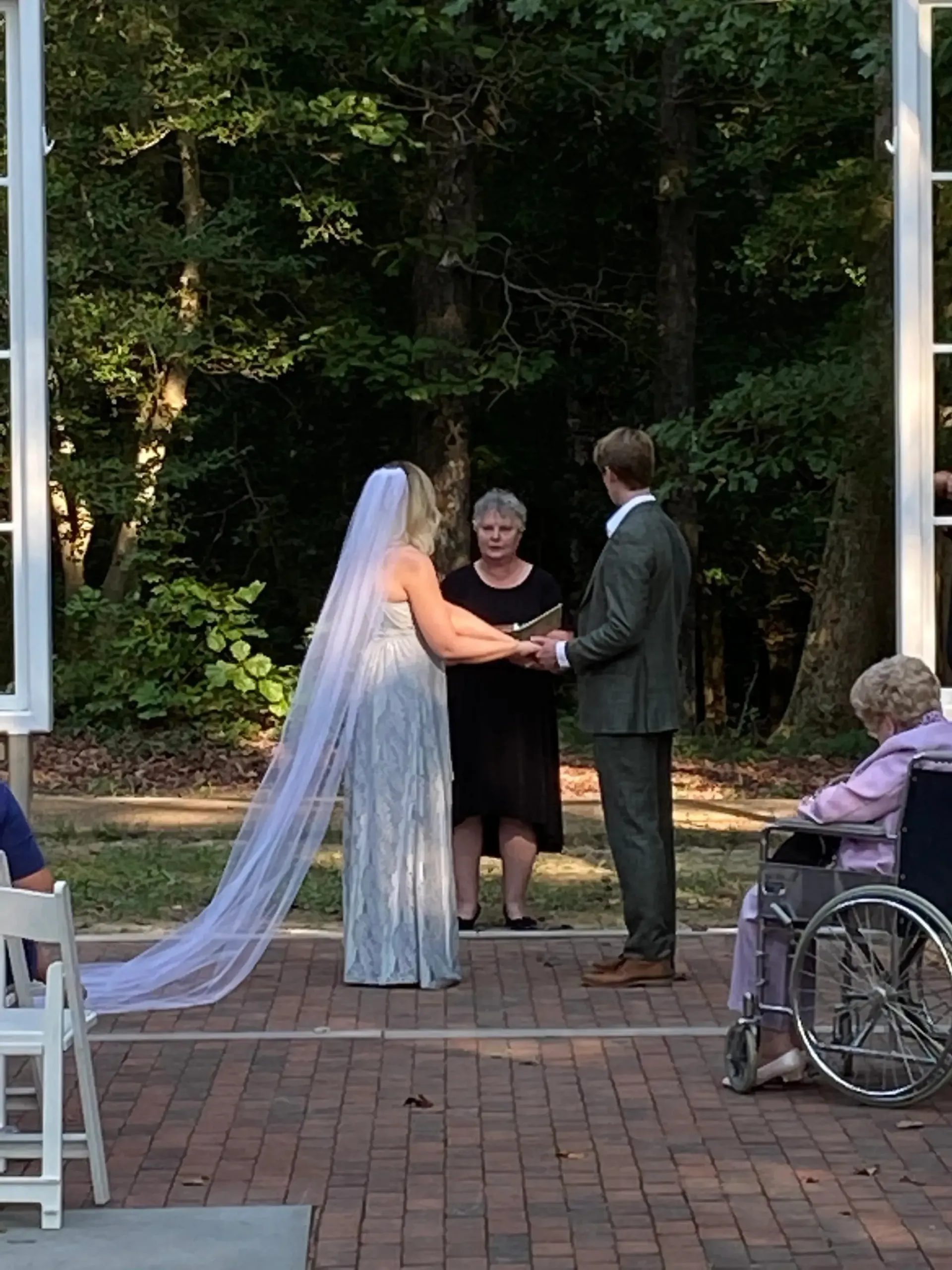 Wedding ceremony outdoors under an arch, bride in white veil holding hands with groom, officiant nearby.