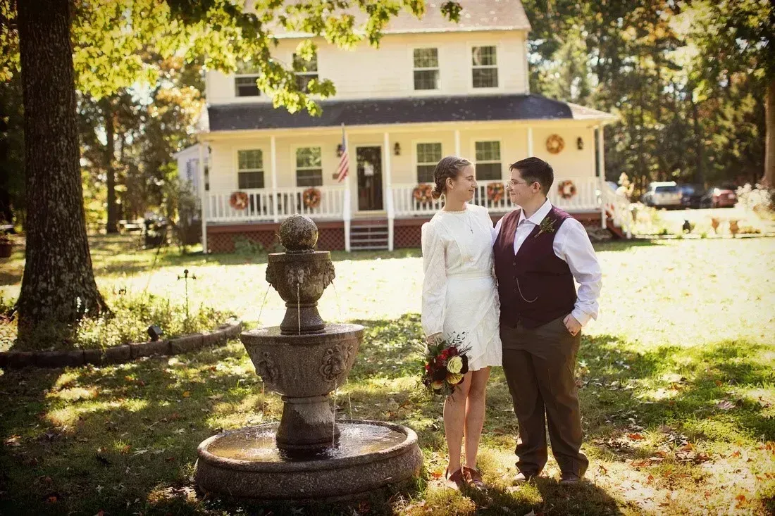 Couple standing together in a sunny yard beside a stone fountain, with a white house in the background