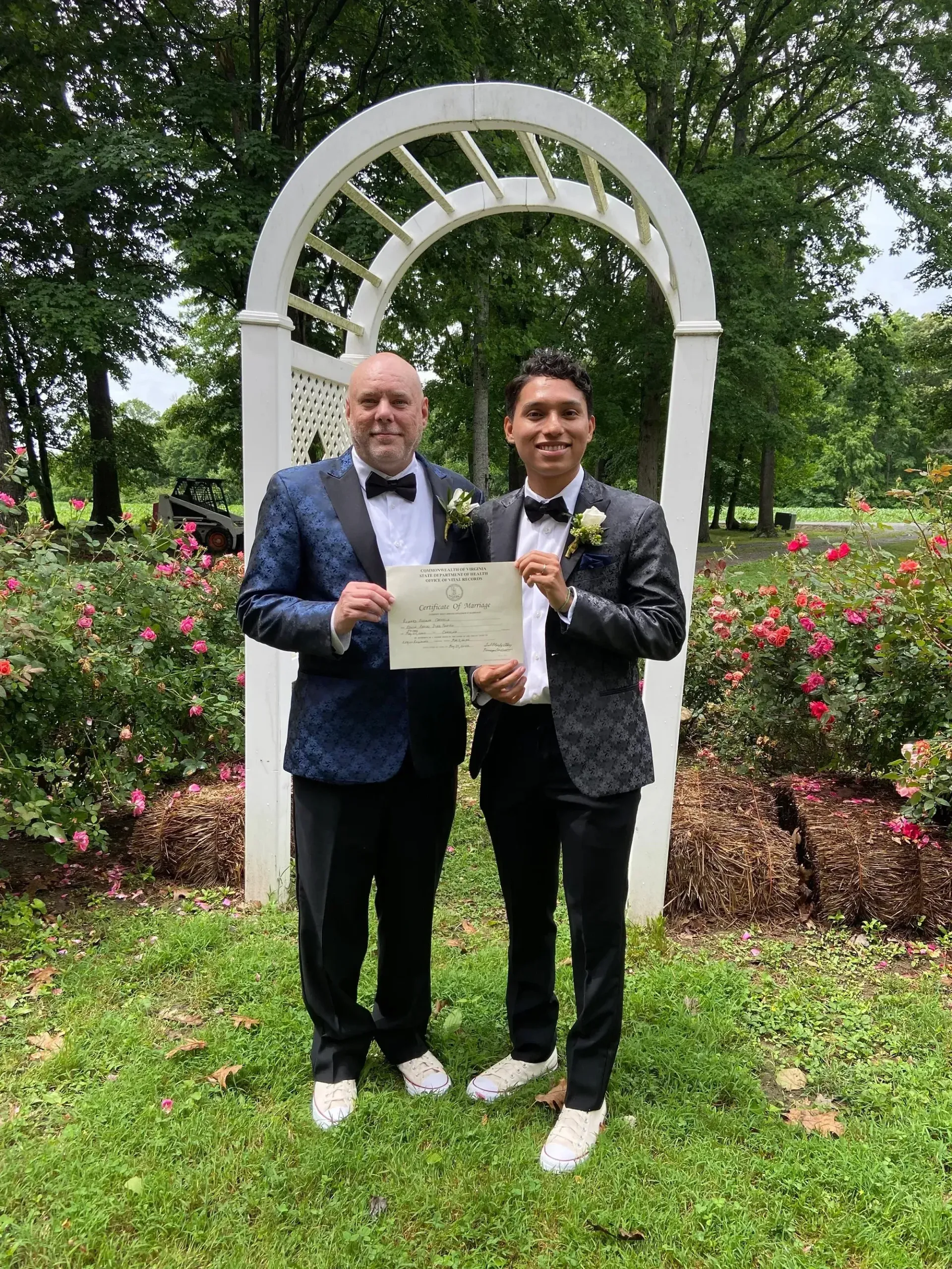 Two men in suits holding a certificate under a white garden arch with flowers.