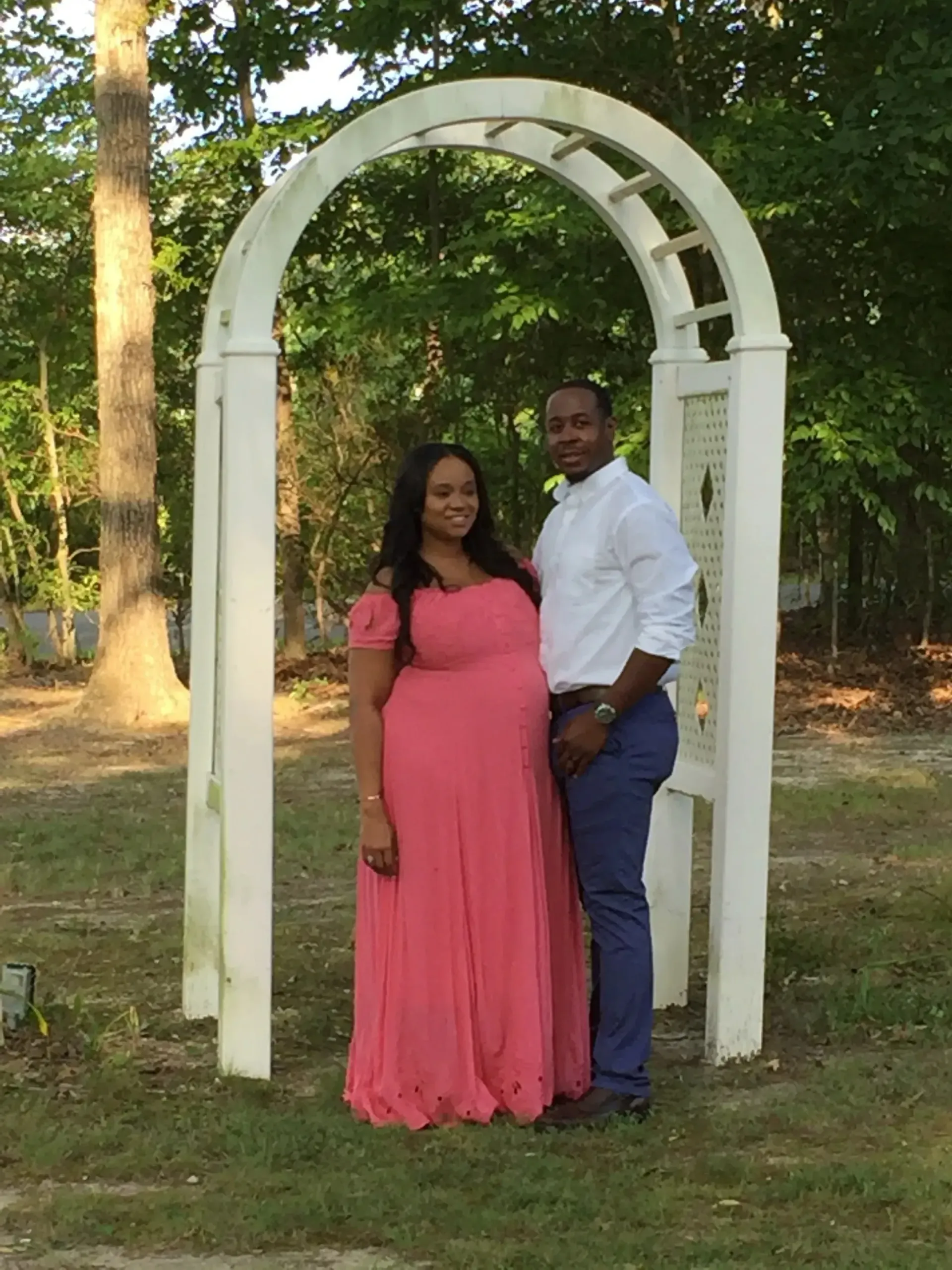 Couple standing under a white garden arch outdoors, with trees and grass in the background.