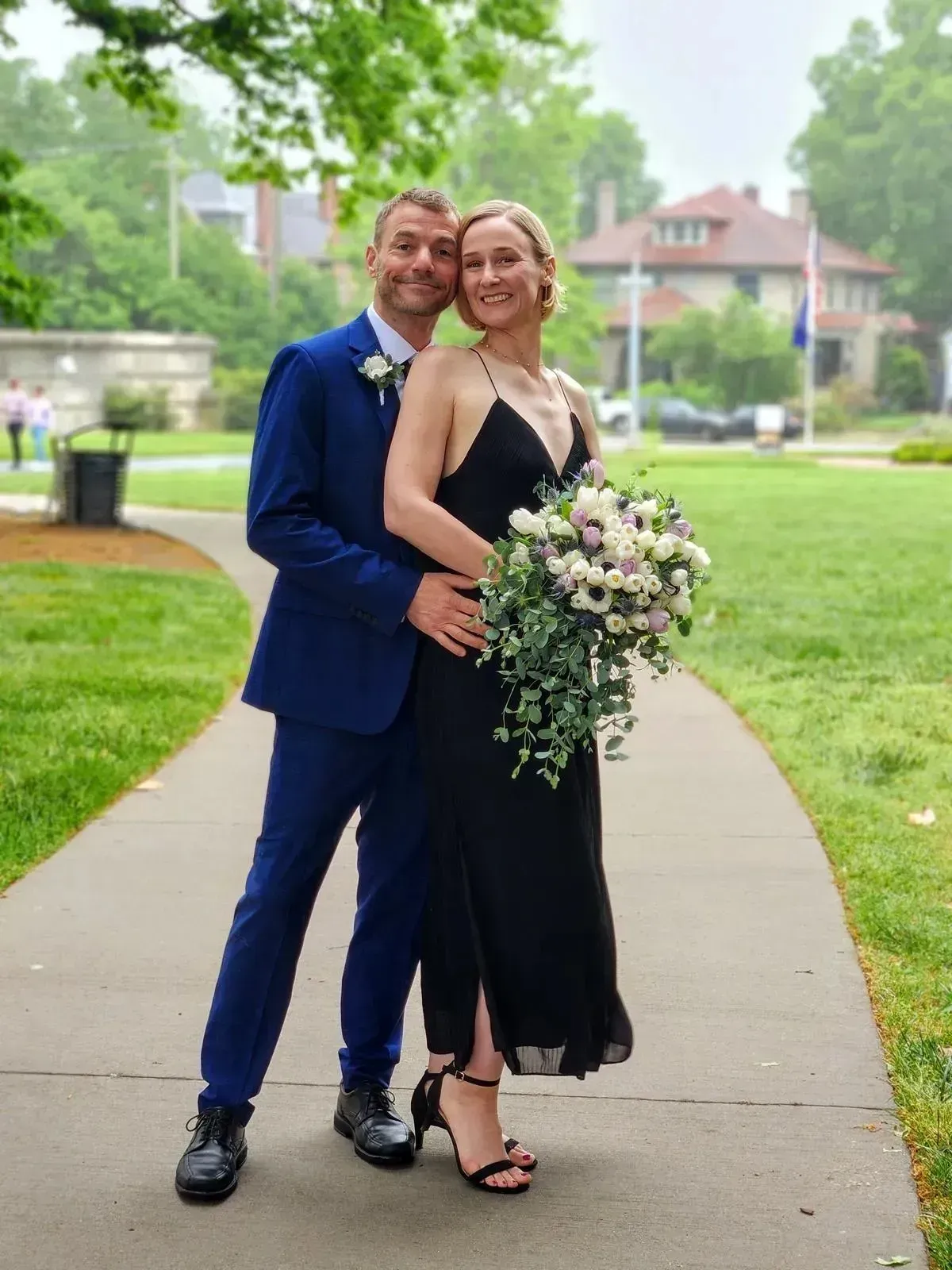 Couple posing on a sidewalk in formal attire, holding a bouquet in a green park.
