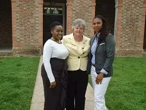 Three women standing together on a lawn in front of a brick building.