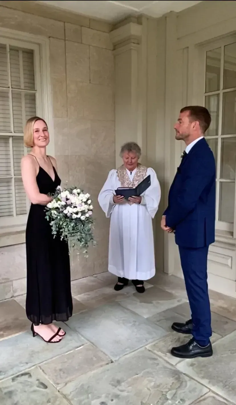 Wedding ceremony with couple facing officiant in a stone-covered entryway, bride holding bouquet in black dress